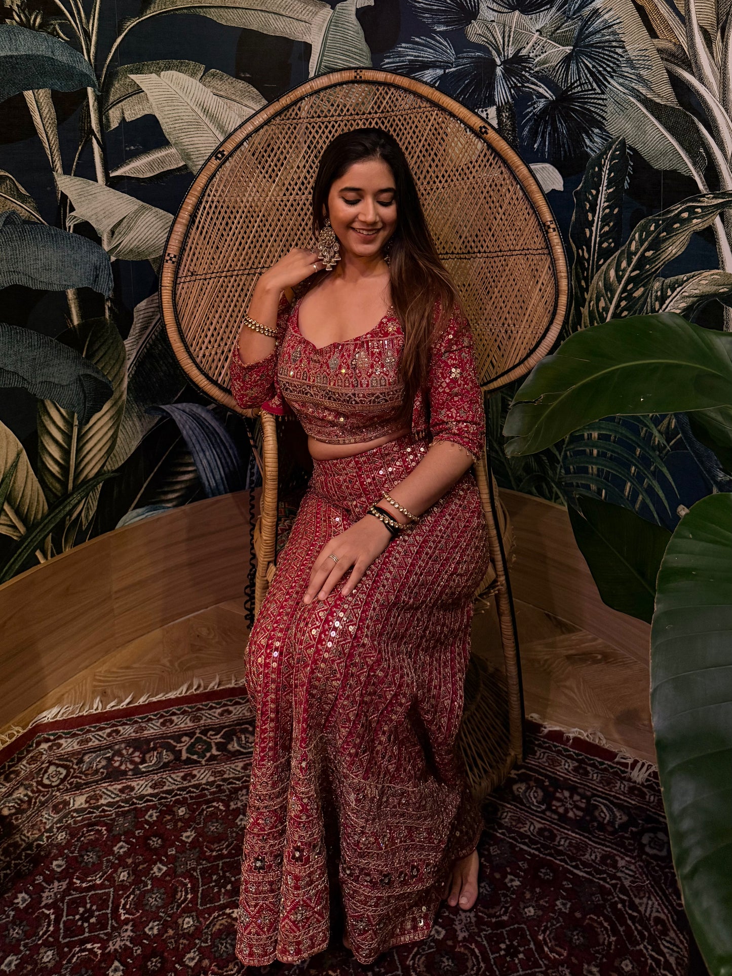 Woman in a red patterned outfit sitting in a decorative chair with a floral wall and rug in the background.