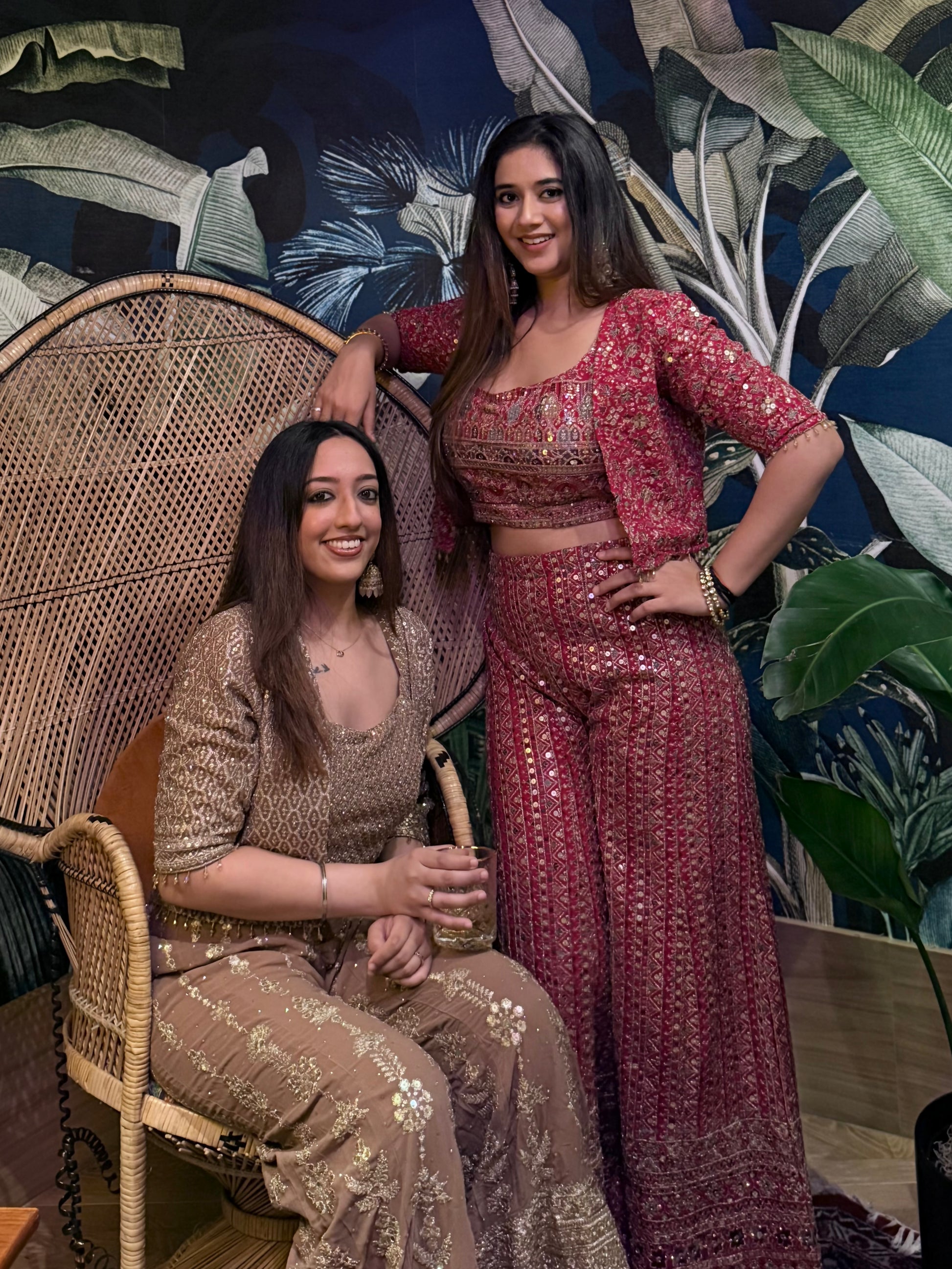 Two women in traditional outfits posing against a decorative wall with plants.